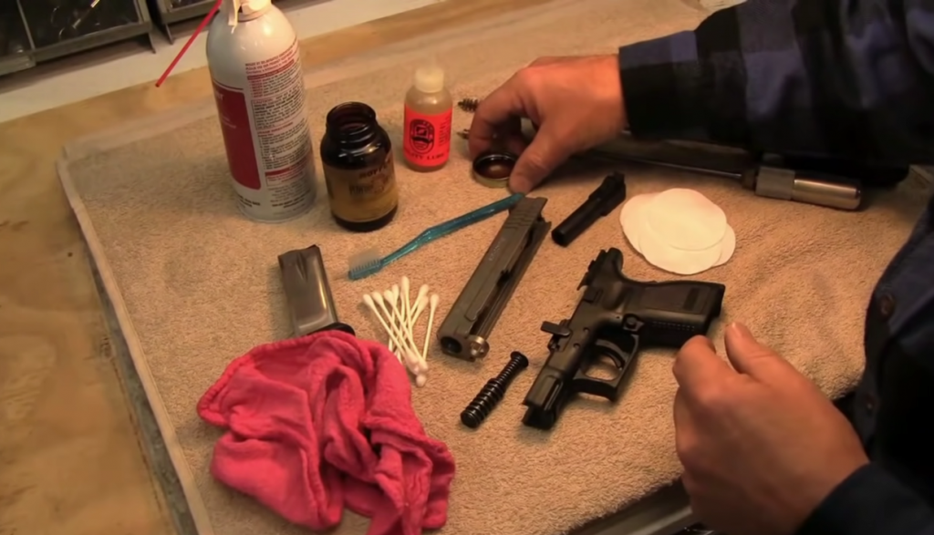 man's hand about to clean a gun with many gun cleaning tools and a disassembled handgun on a table